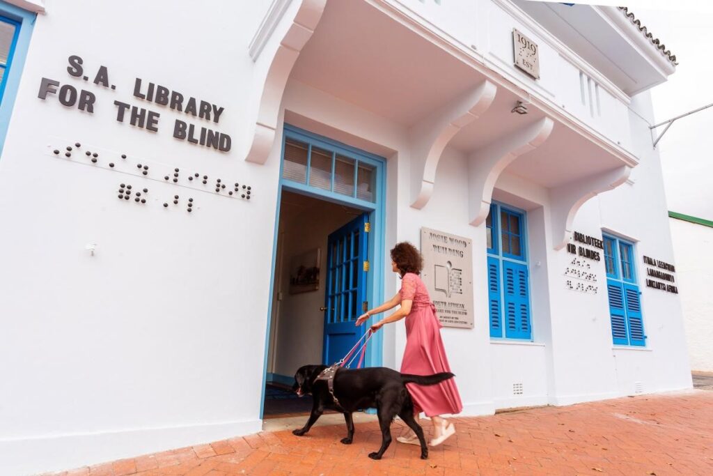 A woman and her guide dog entering the S.A. Library for the blind. A large white building with braille and text. 