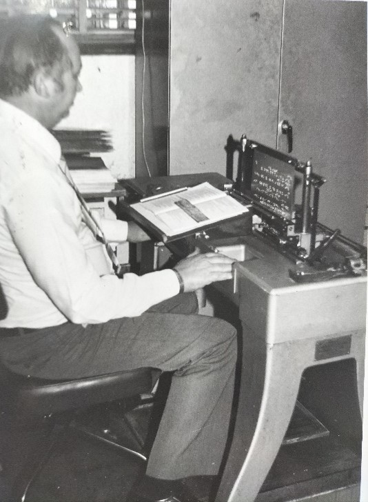 Mr Antoni Zeelie, Braille Services manager from 1963-1996, embossing a braille master plate. He is sitting at the embossers table with a page in front of him.