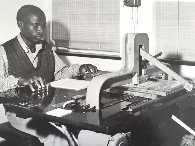 Mr Mandla Ralph Mncube, our first African braille transcriber, brailling a book on the German-manufactured "Marburg Braille Stereotyper" He is feeling the gauge aluminum sheets, there is a slate with a handle to pick up in front of him on the slate as well.