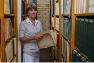 Hester, an elder white female with a braille book open in her hands, Standing between shelves of books.