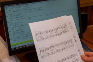 A person is holding a sheet of music in front of a computer.