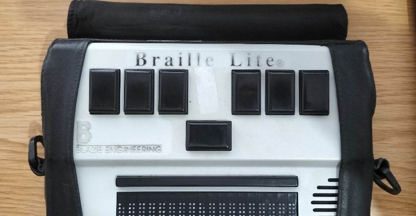 a rectangular device with 7 leys at the top and a bar showing braille below. It has a leather casing.